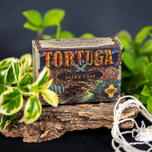 Mydlo-do-brody-Tortuga-110-g-w-opakowaniu-z-kolorowa-grafika.-Widoczny-napis-TORTUGA-z-fantazyjna-czcionka,-informacja-HANDCRAFTED-BEARD-SOAP.-Produkt-lezy-na-kawalku-drewna,-otoczony-zielonymi-liscmi,-na-ciemnym-tle..jpg