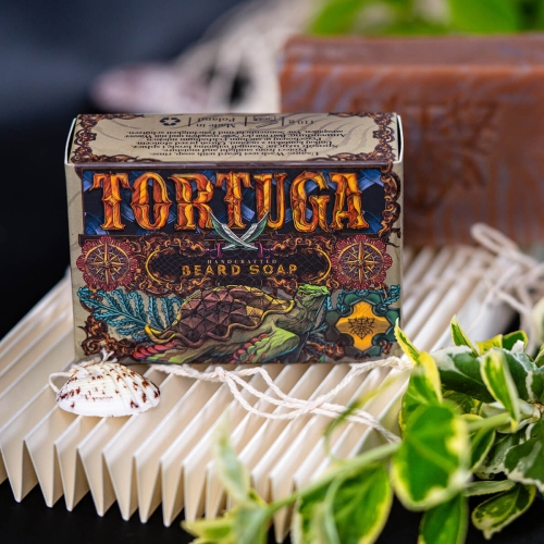 Mydlo-do-brody-Tortuga-110-g-w-ozdobnym-opakowaniu-z-grafika-zolwia-na-brazowym-tle,-widoczne-napisy-“TORTUGA-i-“BEARD-SOAP.-Produkt-lezy-na-falowanej,-jasnej-powierzchni,-w-otoczeniu-roslinnosci-oraz-muszli,-co-nadaje-naturalny-charakter..jpg